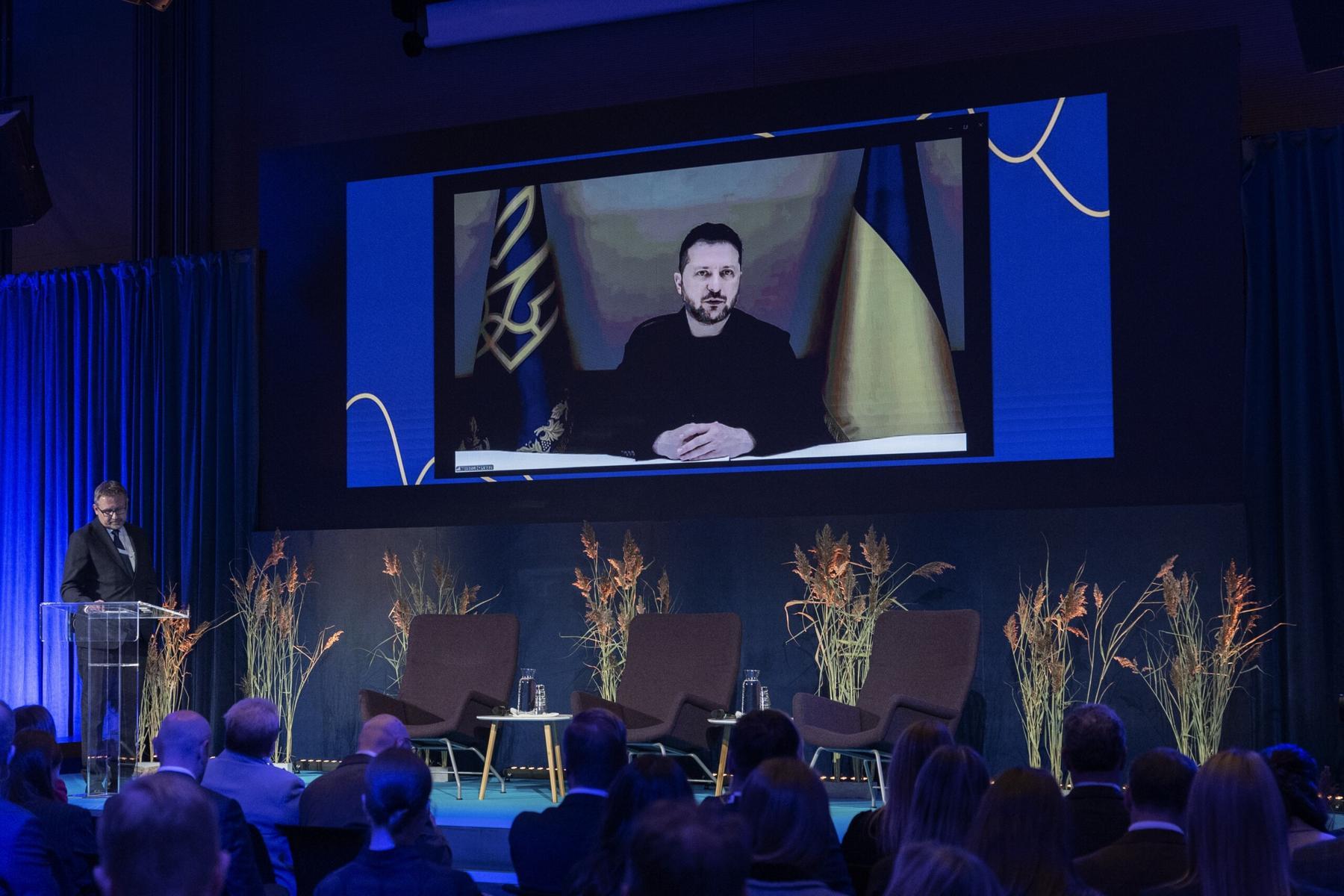 President Zelenskyy on a screen, with the flag of Ukraine behind him.