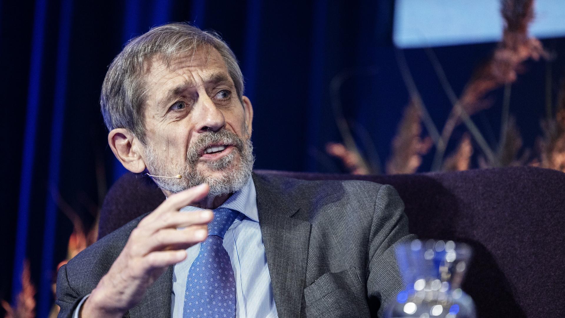 A speaker seated on stage, speaking and gesturing during a panel discussion.
