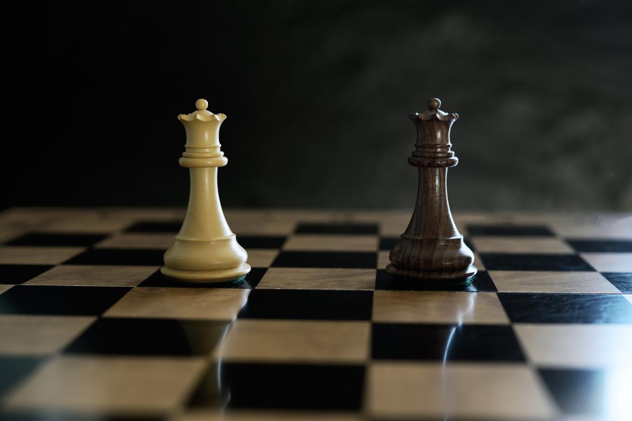 Two opposing chess queen pieces stand facing each other on a black and white chessboard under dramatic lighting.