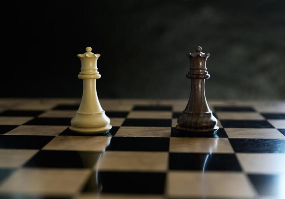 Two opposing chess queen pieces stand facing each other on a black and white chessboard under dramatic lighting.